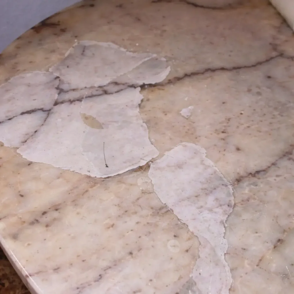 Peeling protective coating flaking off marble table surface exposing raw stone before professional restoration